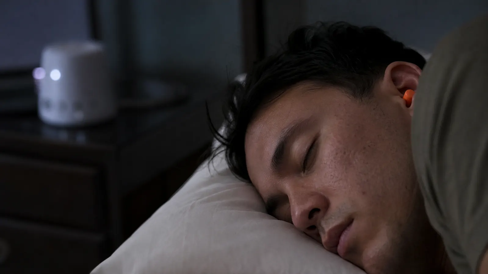 Person sleeping peacefully with foam earplugs in, white noise machine visible on dresser in background