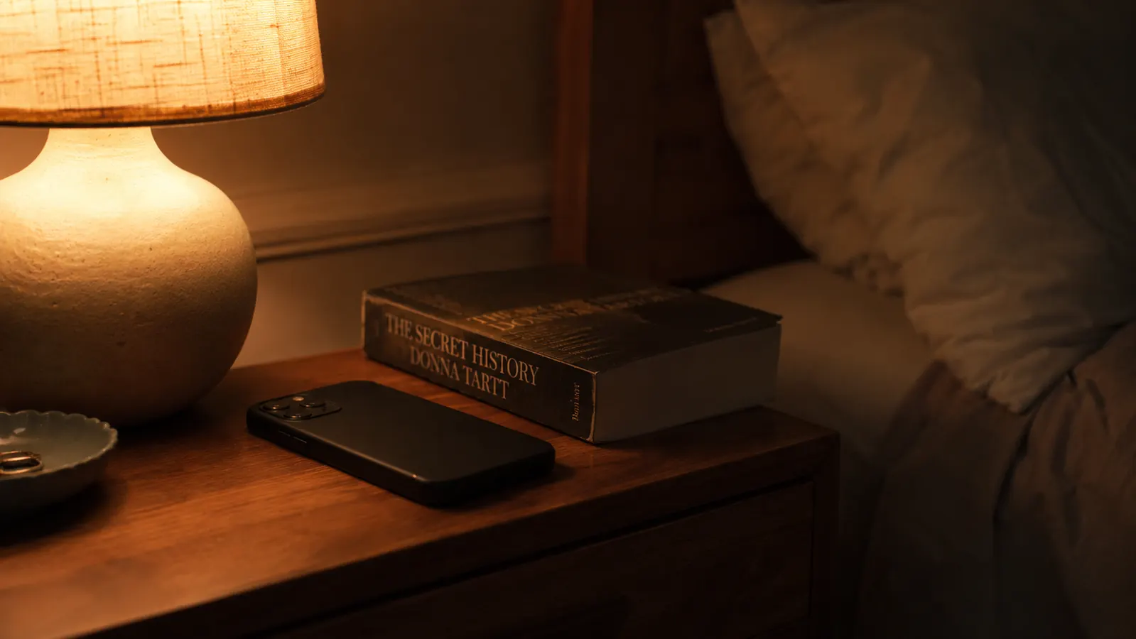 Cozy bedroom at night with a dim warm lamp on, phone face-down on the nightstand, a book nearby