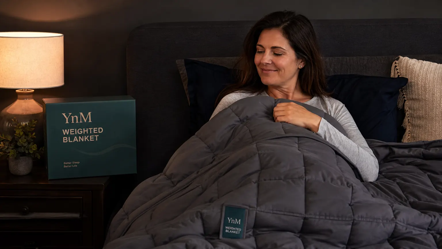 Person pulling a gray YnM weighted blanket up over themselves in bed, glass bead texture visible in the fabric squares