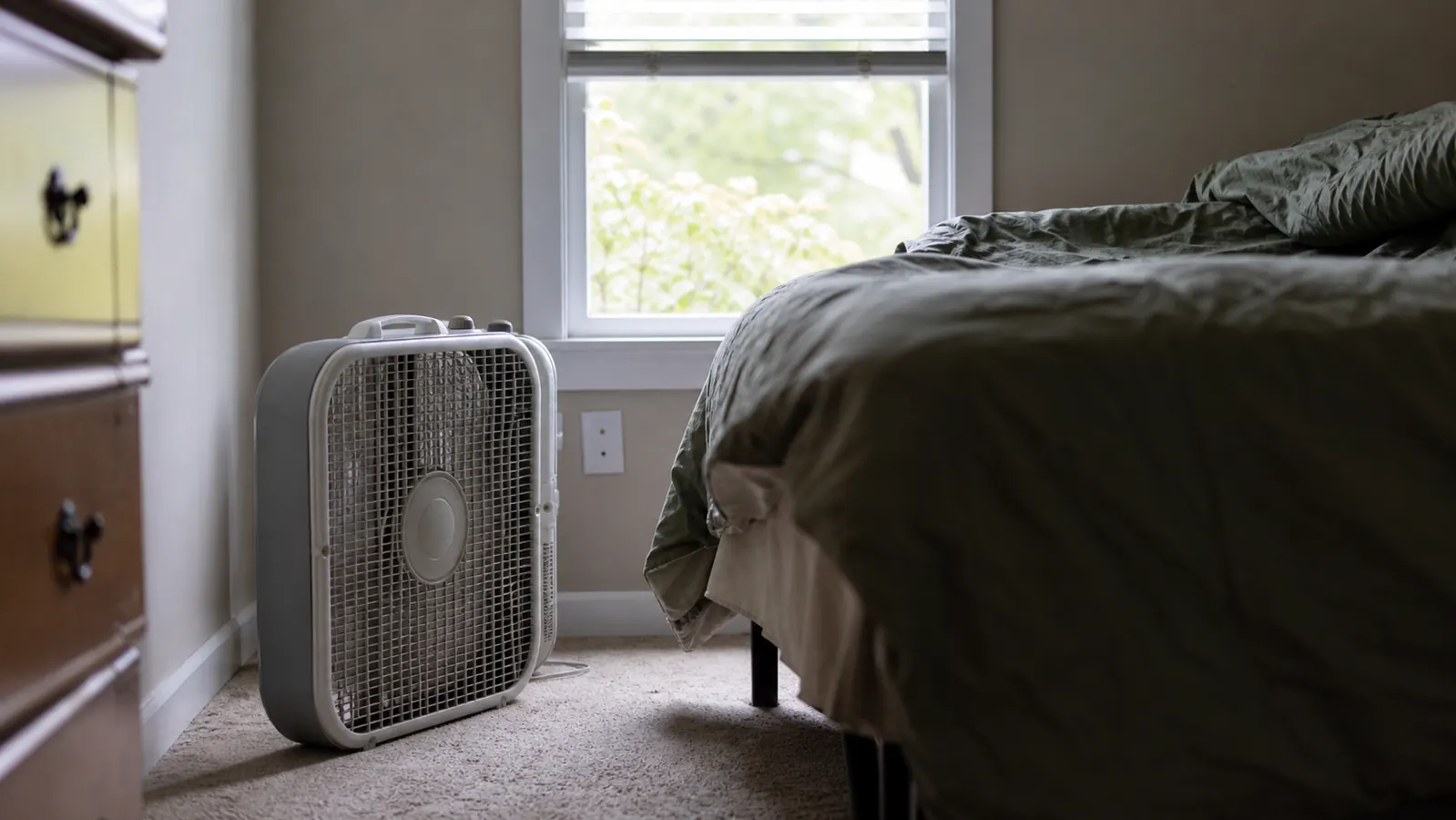Box fan sitting on the floor of a bedroom next to a bed, cord running to outlet